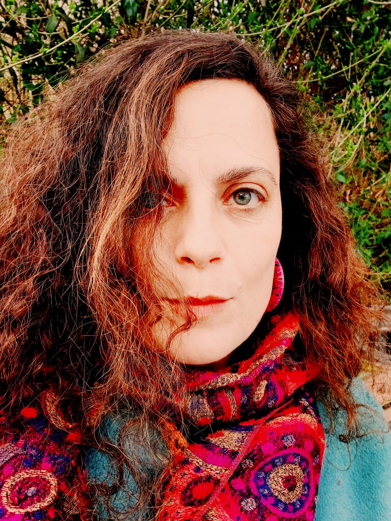 Close-up portrait of a woman with wavy, curly hair wearing a colorful patterned scarf, with a natural background.